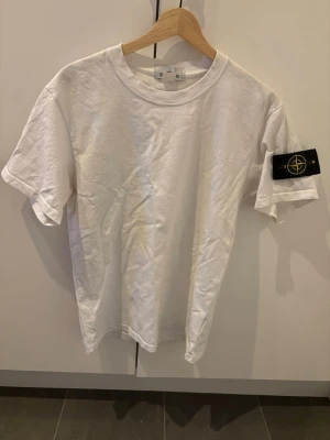 Vit Stone Island t-shirt - Säljer en clean vit t-shirt från Stone Island med klassisk logga på ärmen. T-shirten har rund hals och korta ärmar, perfekt basic-plagg för alla tillfällen. Tillverkad i mjuk bomull för skön känsla och snygg passform.