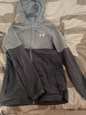 Grå vindjacka från Under Armour - Snygg tvåfärgad vindjacka från Under Armour i ljusgrått och mörkgrått. Jackan har huva, hel dragkedja framtill och två sidofickor. Perfekt för blåsiga dagar och sportiga outfits. Logga på bröstet och tryck på ärmslutet.
