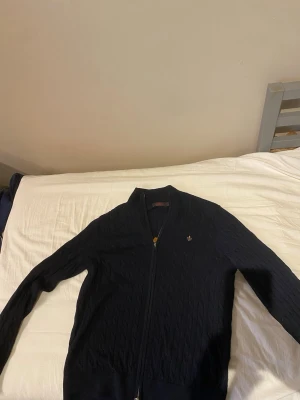 Mörkblå kabelstickad Morris kofta - Snygg mörkblå kabelstickad kofta med dragkedja framtill och liten broderad logga på bröstet. Koftan har ribbade muddar vid ärmslut och nederkant samt en klassisk passform. Perfekt för lager på lager och enkel att matcha. Äkta Morris! Säljs för att den inte används så mycket