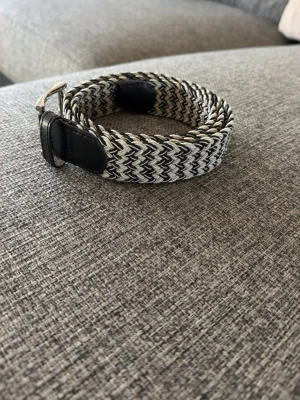 Svartvitt flätat bälte med läderdetaljer - Snyggt svartvitt bälte med zigzag-mönster och flätad design. Bältet har svarta läderdetaljer vid spännet och ändarna, vilket ger en stilren kontrast. Perfekt accessoar för att lyfta din outfit.