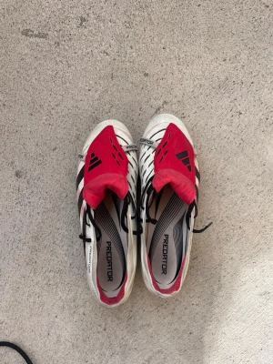 Adidas Predator fotbollsskor röd/vit - Säljer ett par Adidas Predator fotbollsskor i vitt med svarta detaljer och röda tungor. Skorna har klassisk snörning, mönstrad ovandel och dobbar för bra grepp på planen. Perfekta för dig som vill sticka ut på matchen och få grym bollkontroll.