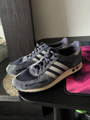 Adidas LA Trainer - Säljer ett par klassiska Adidas LA Trainer sneakers i mörkblått med vita detaljer och de ikoniska tre ränderna på sidan. Ovandelen är i mesh och mocka, och sulan har de typiska pluggarna. Perfekta för dig som gillar retrostil och sportig look.