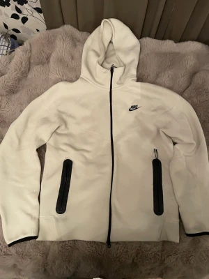 Vit Nike hoodie med dragkedja - Snygg vit hoodie från Nike med svart dragkedja och svarta detaljer på fickorna. Klassisk Nike-logga på bröstet och huva för extra stilpoäng. Tillverkad i mjukt material som känns skönt mot huden. Perfekt för dig som gillar sportig och clean look. Den har ett litet märke men inget som man tänker på. 