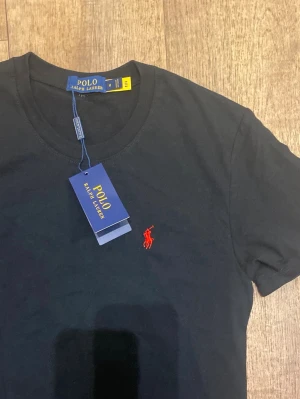 Svart t-shirt från Polo Ralph Lauren (Helt ny / Oanvänd) - Klassisk svart t-shirt från Ralph Lauren med röd broderad logga på bröstet. T-shirten har rund halsringning och korta ärmar. Tillverkad i mjuk bomull för en skön känsla och enkel stil. Storlek M men passar mer som Small. Snabb Leverans!