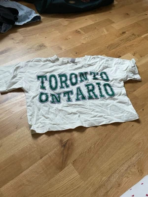 Vit croppad t-shirt Toronto Ontario - Säljer en vit croppad t-shirt från Lindex med grön text 'Toronto Ontario' på framsidan. T-shirten har korta ärmar och är gjord i mjuk bomull, perfekt för en chill och avslappnad stil.