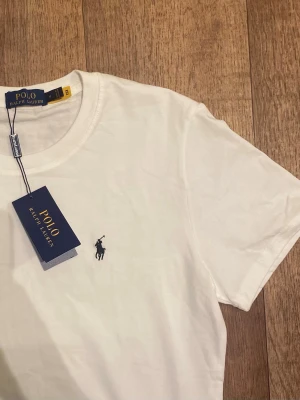 Vit t-shirt från Polo Ralph Lauren (Helt ny / Oanvänd) - Klassisk vit t-shirt från Ralph Lauren med den ikoniska broderade loggan på bröstet. T-shirten har rund halsringning och korta ärmar. Tillverkad i mjuk bomull för en skön och avslappnad känsla. Perfekt basplagg till garderoben. Storlek medium men passar mer som small. Snabb Leverans!