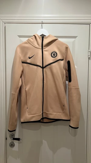 Beige Chelsea Nike hoodie - Snygg beige hoodie från Nike med Chelsea FC-logga på bröstet och svart Nike-märke. Dragkedja framtill, svarta detaljer och fickor på sidorna samt en extra ficka på ärmen. Tillverkad i mjukt material och har huva för extra stilpoäng. Storleken är S men passar även M.