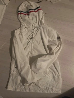 Vit vindjacka från Moncler - Snygg vit vindjacka från Moncler med huva och dragkedja framtill. Jackan har Moncler-logga på ärmen och detaljer med röda och marinblå ränder på huvan. Tillverkad i lätt material, perfekt för blåsiga dagar. Praktiska fickor med knappstängning och justerbar dragsko i huvan.
