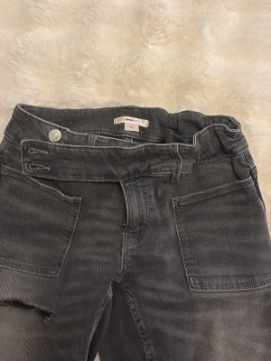 Svarta jeans från Gina Tricot - Säljer ett par svarta jeans från Gina Tricot med raka ben och slitning vid knät. Jeansen har stora framfickor och dubbla knappar i midjan. Materialet är klassisk denim med en tvättad look som ger en cool vibe.