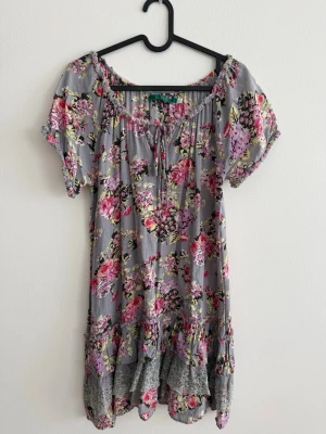 RoseBud Tunika/Kort Klänning - Fin och luftig tunika/kort klänning från Rose Bud med romantiskt blommönster i rosa, grått och beige. Modellen har en lös passform med korta ärmar, knytning framtill vid halsen och fina detaljer med volang och spets nedtill.  Kan bäras både som kort klänning eller tunika över t.ex. jeans eller shorts.  Märke: Rose Bud Storlek: S Längd: ca 70 cm