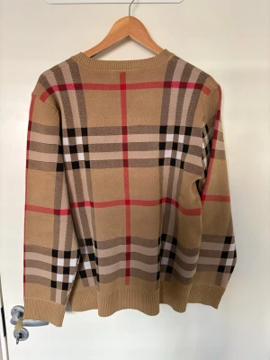 Beige rutig stickad tröja Burberry - Säljer en beige stickad tröja från Burberry med det klassiska rutmönstret i svart, vitt och rött. Tröjan har rund halsringning, långa ärmar och ribbade muddar vid ärmslut och nederkant. Perfekt för dig som gillar tidlös och ikonisk stil.