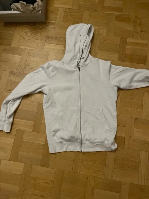Vit zip hoodie med huva - En enkel och stilren vit hoodie med dragkedja framtill och två stora fickor. Hoodien har en klassisk huva med snörning och ribbade muddar vid ärmar och nederkant. Perfekt för en avslappnad och bekväm look.