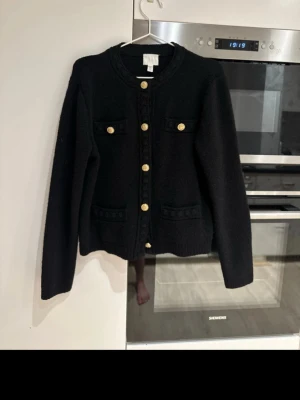 Svart kofta med guldknappar - Stilren svart kofta med rund hals och dekorativa guldknappar framtill. Fyra fickor med knappdetaljer ger en klassisk look. Koftan är stickad och har långa ärmar, perfekt för lager på lager. Passar till både jeans och kjol för en chic outfit. Nypris 499