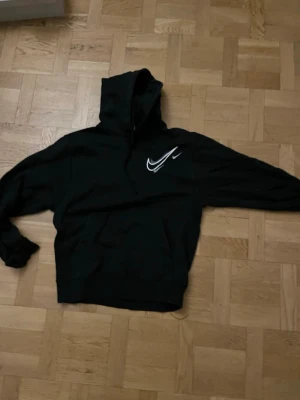 Svart hoodie från Nike med vit logga - Säljer en svart hoodie från Nike med stor vit Swoosh-logga på bröstet. Klassiskkänguruficka framtill och huva med dragsko. Perfekt för en sportig och avslappnad stil.