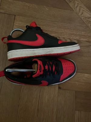 Nike svart-röda sneakers lows - Säljer ett par snygga Nike sneakers i svart och rött med klassisk låg siluett. Skorna har svart ovandel med röda detaljer, röd sula och Nike-logga på sidan och plös. Perfekta för dig som gillar streetwear och vill sticka ut.