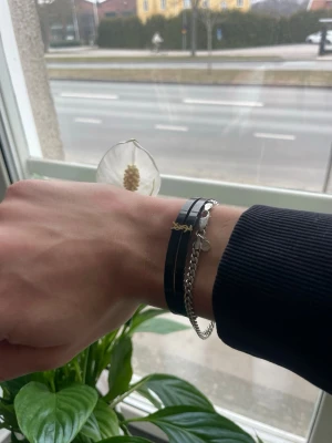 Snyggt armband - Snyggt svart läderarmband från Yves Saint Laurent med guldfärgad YSL-logga och dubbla remmar. Kombineras med en stilren silverfärgad kedja för en modern look. Perfekt accessoar för dig som gillar exklusiva detaljer och trendig design.