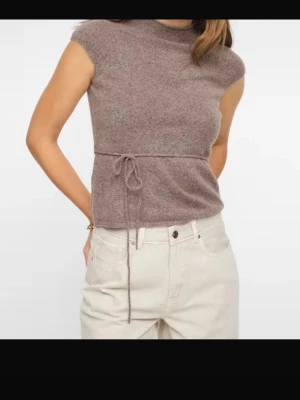 Brun ärmlös topp med knyt - Mjuk brun topp med hög hals och ärmlös design. Midjan markeras av ett tunt knytband som ger en snygg siluett. Perfekt att styla med både jeans och kjol för en trendig look.