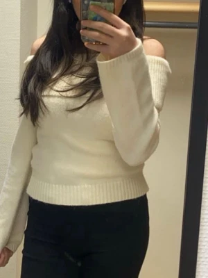 Off shoulder vit stickad tröja - Trendig off shoulder stickad tröja i krämvit färg. Tröjan har långa ärmar, bred ribbad mudd vid midjan och är tillverkad i ett mjukt, tjockt material. Perfekt för dig som vill ha en stilren och modern look.