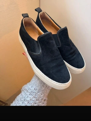 Svarta slip-on sneakers i mocka/läder - Stilrena svarta slip-on sneakers i mocka med skinndetaljer, vit sula och elastiska paneler på sidorna. Skorna har en enkel och clean design med dragflikar bak för enkel påtagning. Sparsamt använda men väldigt bekväma tack vare ortopedisk sula i läder. Angiven storlek är 38 men dem passar snarare en 39a.