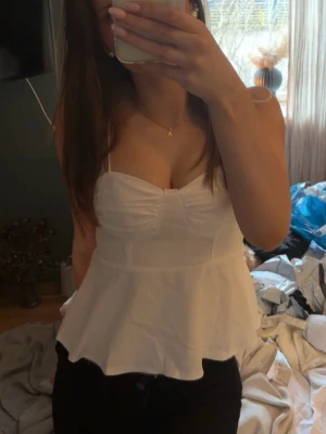 Söt peplum- topp  - Säljer en söt vit linnetopp med smala axelband och volang nedtill. Toppen har rynkdetalj framtill och smockad rygg för skön passform. Perfekt för varma dagar och enkel att styla med jeans eller kjol. Helt ny!