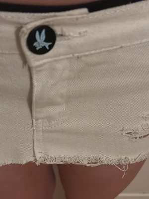 Jeanskjol från oneteaspoon - Säljer en beige kort jeanskjol med rå, fransig kant och slitna detaljer. Kjolen har en knapp med en fågel på framtill och klassisk femficksdesign. Perfekt för dig som gillar en avslappnad och trendig stil.