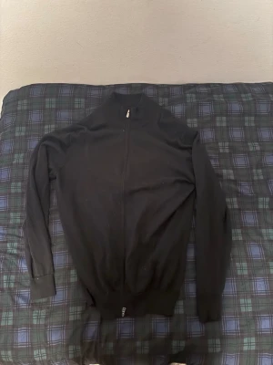 Svart stickad ziptröja från Boggi - Snygg svart stickad tröja från Boggi med dragkedja framtill och hög krage. Tröjan har långa ärmar och ribbade muddar vid ärmslut och nederkant. Perfekt för lager på lager och stilrena outfits.