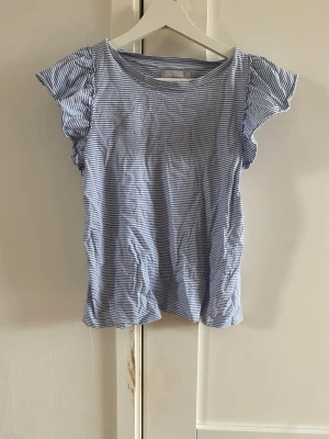 Blåvit randig topp från Tiki - Söt blåvit randig topp från Tiki med korta, lätt volangiga ärmar och rund halsringning. Tillverkad i mjuk bomull som känns skön mot huden. Perfekt för en avslappnad och fräsch look.