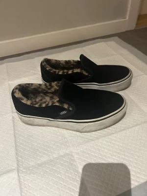 Svarta Vans slip-on i mocka med foder - Svarta slip-on skor från Vans med fluffigt leopardmönstrat foder. Yttermaterialet är i mockaliknande tyg och sulan är vit med klassisk Vans-logga på sidan. Använda ett fåtal gånger men superkomfortabla. 