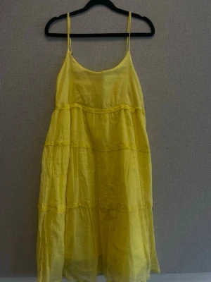 Gul långklänning med tunna axelband - Luftig gul långklänning med tunna axelband och volangdetaljer. Klänningen har en lös passform och är tillverkad i ett lätt, skrynkligt tyg som passar perfekt till varma dagar.