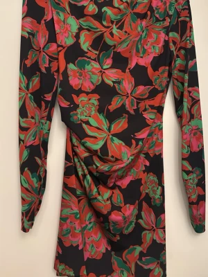 Blommig långärmad kortklänning - Snygg kort klänning med svart bas och stora blommiga mönster i rosa, rött och grönt. Klänningen har långa ärmar och en draperad detalj framtill som ger en cool siluett. Perfekt för dig som vill sticka ut med färg och mönster.zara 