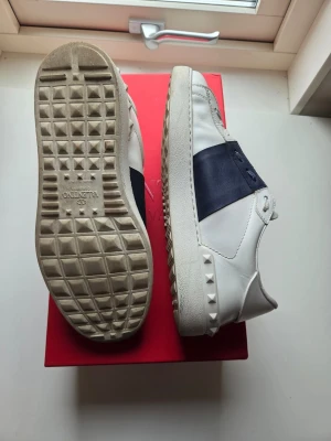 Valentino vita sneakers med blå detalj - Snygga vita sneakers från Valentino med bred mörkblå rem över ovandelen och coola nitar längs sulkanten. Skorna är låga och har en klassisk siluett med vita snören och gummisula. Perfekta för dig som vill ha en stilren men edgy look. Skorna har lite skav i hälen av mycket användning vilket inte syns eller känns när man har de på sig.