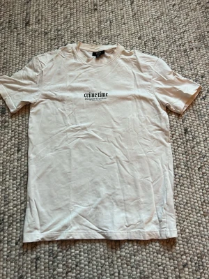 Vit t-shirt Crime Time tryck - Vit t-shirt från New Yorker med svart texttryck på bröstet och stort, detaljerat tryck på ryggen med olika texter och grafik. T-shirten har rund hals och korta ärmar. Materialet är mjukt bomullstyg och modellen är rak och avslappnad.