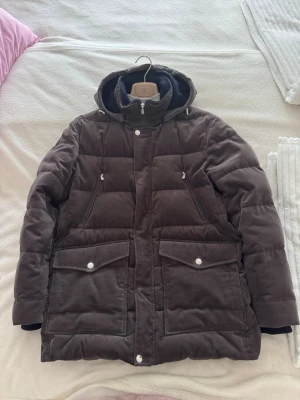 Brunello Cucinelli brun pufferjacka - Säljer en brun pufferjacka från Brunello Cucinelli i mjuk manchester. Jackan har huva, dragkedja och tryckknappar framtill, samt stora fickor med lock och vita knappar. Perfekt för kalla vinterdagar. Retail pris: 60k plus