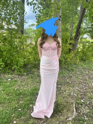 Ljusrosa långklänning med spetsdetaljer - Elegant ljusrosa långklänning med tunna axelband, v-ringad urringning och vackra spets- och broderidetaljer över bysten och ena sidan. Klänningen har figurnära passform och är tillverkad i glansigt satinliknande material. Perfekt för bal eller student. 