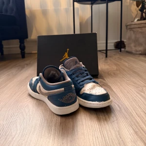 Nike Air Jordan 1 Low blå/vit - Nike Air Jordan 1 Low sneakers i blått och vitt med grå detaljer. Klassisk Jordan-logga på hälen och Swoosh på sidan. Skorna har snörning, rund tå och platt sula. Tillverkade i skinn och syntetmaterial, med meshfoder för extra komfort.