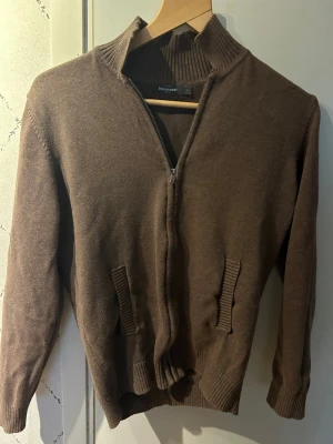Brun stickad cardigan från dressmann - Mysig brun stickad cardigan från dressmann med hög krage och ribbade muddar. Tröjan har två fickor framtill och dragkedja hela vägen. Perfekt för lager på lager och enkel att matcha med olika outfits.
