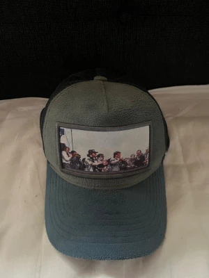 Grå meshkeps med motiv framtill - Säljer en grå keps med mesh baktill och justerbart spänne. Framtill finns ett tygmärke med ett tryckt motiv av personer på motorcyklar. Kepsen har böjd skärm och klassisk trucker-modell. Perfekt för dig som vill ha en unik accessoar.