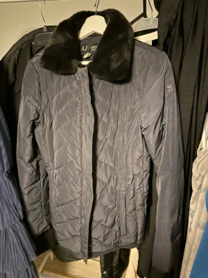 Mörkblå kappa från Armani Jeans - Stilren mörkblå kappa från Armani Jeans med mjuk, svart fuskpälskrage. Kappan har diskreta fickor och ett quiltat mönster som ger en modern look. Perfekt för kyliga dagar när du vill vara både varm och snygg.