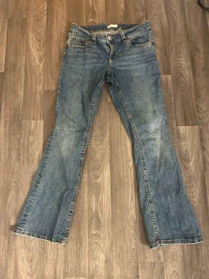 Blå bootcut jeans med fickdetaljer - Ett par klassiska blå bootcut jeans med fem fickor och snygga sömmar. Jeansen har knapp- och dragkedjestängning samt dekorativa ficklock med knappar bak. Perfekta för en avslappnad och trendig look.
