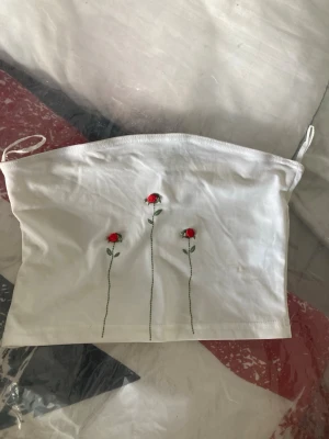 Vit tubtopp med broderade rosor - Söt vit tubtopp med tre broderade röda rosor framtill. Toppen är axelbandslös och har en enkel, rak passform. Perfekt att matcha med jeans eller kjol för en trendig look.