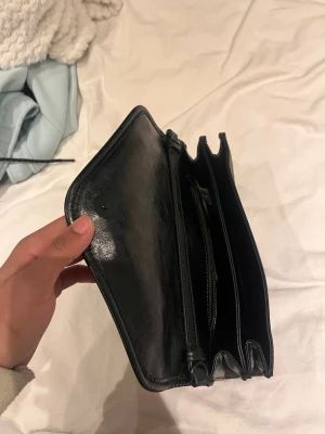 Svart lång plånbok i skinn - Stilren svart plånbok i äkta skinn med flera fack för kort, sedlar och mynt. Klassisk lång modell med tryckknappsstängning och enkel, tidlös design. Perfekt för dig som vill ha ordning och reda på dina grejer.