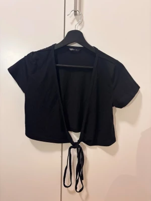 Svart croppad topp med knyt - Trendig svart croppad topp med korta ärmar och knyt framtill. Perfekt att styla över ett linne eller topp för en cool lager-på-lager-look. Enkel och stilren design som passar till många outfits.