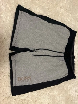 Grå och svarta shorts från Hugo Boss - Snygga shorts från Hugo Boss i grått och svart med logga på vänster ben. De har snörning i midjan och är tillverkade i mjuk bomull, perfekta för en avslappnad stil. Shortsen har en loose passform och praktiska fickor på sidorna.