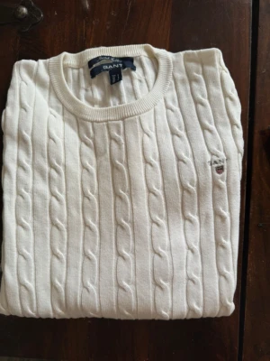 Vit kabelstickad tröja från GANT - Kabelstickad vit tröja från GANT i stretchig bomull. Klassisk rund halsringning och diskret GANT-logga broderad på bröstet. Perfekt för en clean och stilren look. Passar till jeans eller kjol för en snygg vardagsstil.