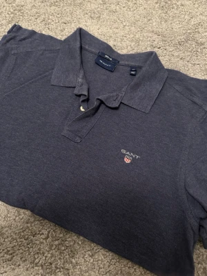 Mörkblå piké från GANT - Snygg mörkblå pikétröja från GANT med klassisk krage och knappar framtill. Tröjan har GANT-logga och sköld broderad på bröstet. Tillverkad i mjuk bomull för en bekväm känsla och passar perfekt till många olika stilar.