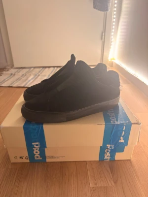 Arigato clean 90 laceless - Snygga helsvarta slip-on sneakers med låg profil och minimalistisk design. Skorna har en ovandel i mockaliknande material och en matchande svart sula. Perfekta för dig som gillar stilrena och enkla sneakers till vardagslooken.