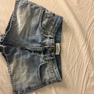 Blå denimshorts från Pull & Bear - Säljer ett par klassiska blå denimshorts från Pull and bear med fickor, bälteshällor och knappgylf. Shortsen har en avslappnad passform och är perfekta för varma dagar. Snygg tvättad look och tidlös design.