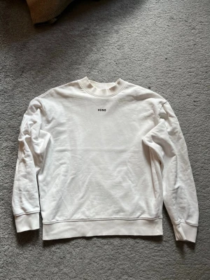 Vit sweatshirt från Hugo Boss - Säljer en stilren vit sweatshirt från Hugo med diskret logga på bröstet. Tröjan har rund halsringning, långa ärmar och ribbade muddar vid ärmslut och nederkant. Perfekt för en clean och enkel look.