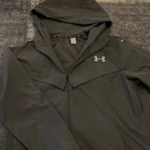 Grå vindjacka från Under Armour - Snygg grå vindjacka från Under Armour med huva och dragkedja framtill. Jackan har en minimalistisk design, diskret logga på bröstet och är tillverkad i lätt, vindtätt material. Perfekt för löprundan eller när du vill ha en sportig look.