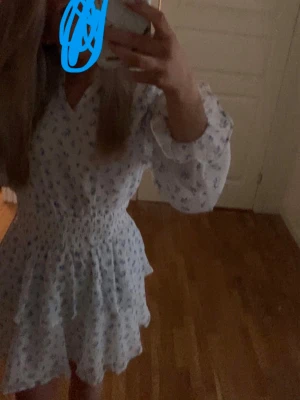 Vit blommig kortklänning med volang - Supersöt vit kortklänning med blått blommönster och volangdetaljer. Klänningen har långärmade, lätt transparenta ärmar och smock i midjan för en bekväm passform. Perfekt för sommarens alla tillfällen!